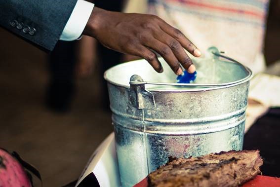 A person in a suit reaches out to pick a blue object from a metallic bucket. A piece of brown bread or cake is in the foreground. The setting appears casual.