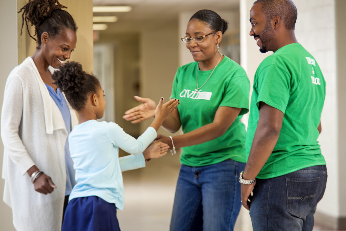 A woman and a young girl exchange high-fives in a bright hallway. Another woman and a man, both in matching green shirts, stand nearby, smiling as they observe the cheerful interaction.
