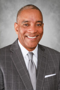 A man in a gray plaid suit and tie smiles warmly at the camera. The background is a plain, light gray, enhancing the professional appearance.