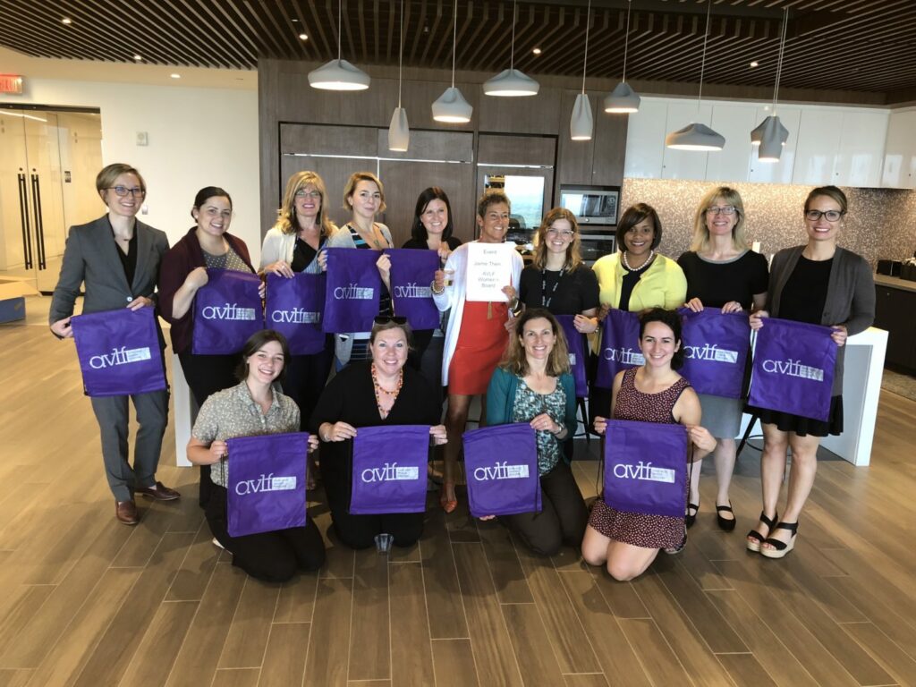 A group of twelve people, mostly women, standing in an office kitchen area. They are smiling and holding purple tote bags with the "CVIH" logo. One person in the center holds a certificate. The room has modern lighting and wooden flooring.