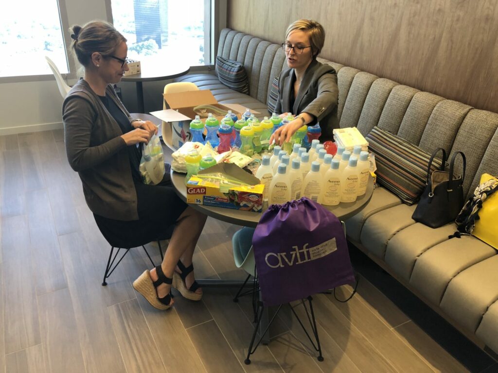 Two people sit at a table filled with baby bottles, sippy cups, and other supplies. They appear to be organizing or packing the items. The room has a sofa and large windows in the background.
