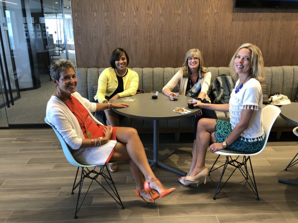 Four women sit around a table in a modern office lounge, smiling. They are dressed in business casual attire, and each has a drink in front of them. The space features a wooden wall and glass partitions in the background.