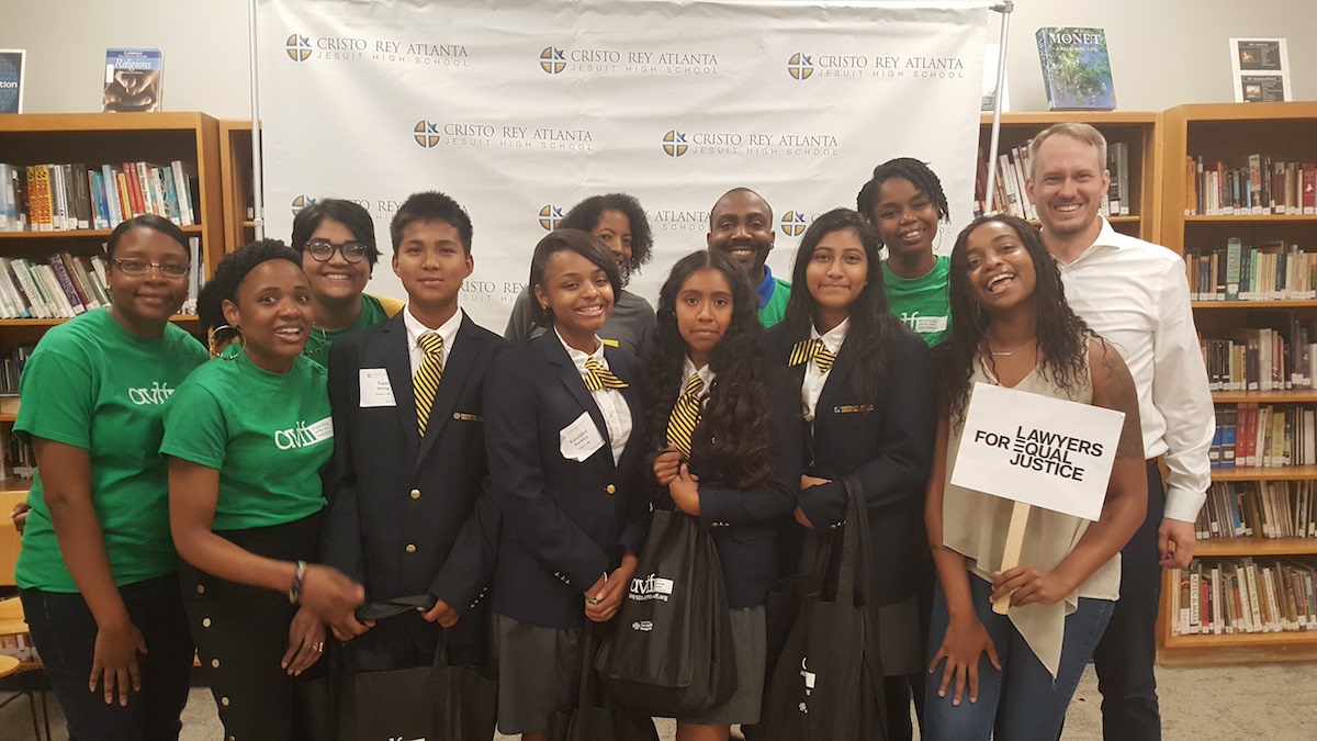 A group of students in school uniforms and adults in green shirts are smiling and standing together in a library. One person holds a sign that reads "Lawyers for Equal Justice." A banner in the background reads "Cristo Rey Atlanta Jesuit High School.
