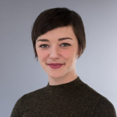 A person with short dark hair and a nose piercing smiles softly at the camera, wearing a dark sweater. The background is a simple gray gradient.
