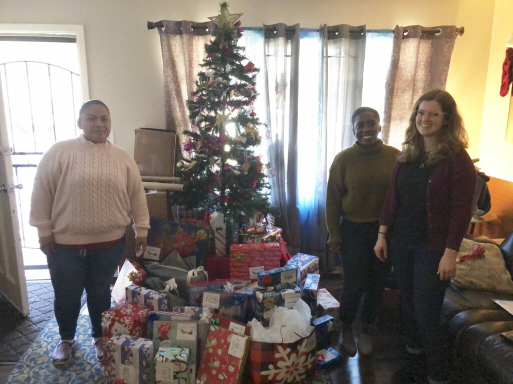 Three people stand smiling beside a decorated Christmas tree with a star on top. Numerous wrapped gifts are piled under the tree. The room is softly lit, and curtains partially cover the windows in the background.
