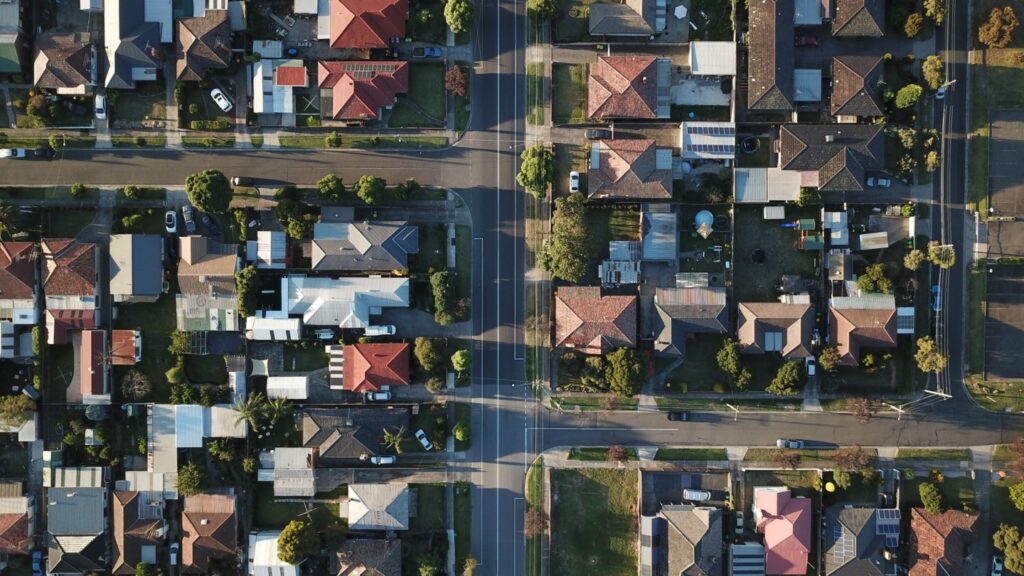 Aerial view of a suburban neighborhood with rows of houses, each with distinct red or gray roofs, separated by streets and lined with trees. Roads intersect in the center, forming a cross pattern. Numerous cars are parked in driveways and along the streets.