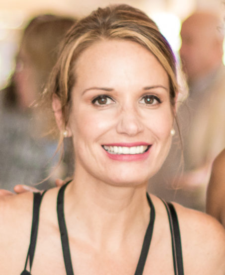 A woman with light brown hair pulled back, smiling at the camera. She is wearing a black tank top and has pearl earrings. The background is softly blurred with people and lights.