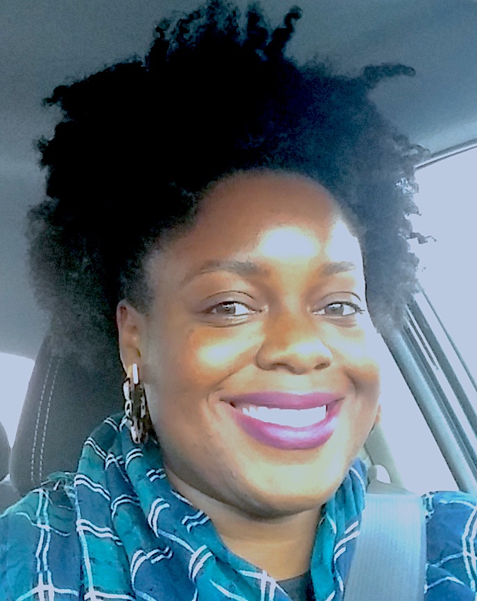 A person with curly hair smiles in a selfie taken inside a car. They are wearing a blue and white patterned scarf and earrings, with sunlight illuminating their face.