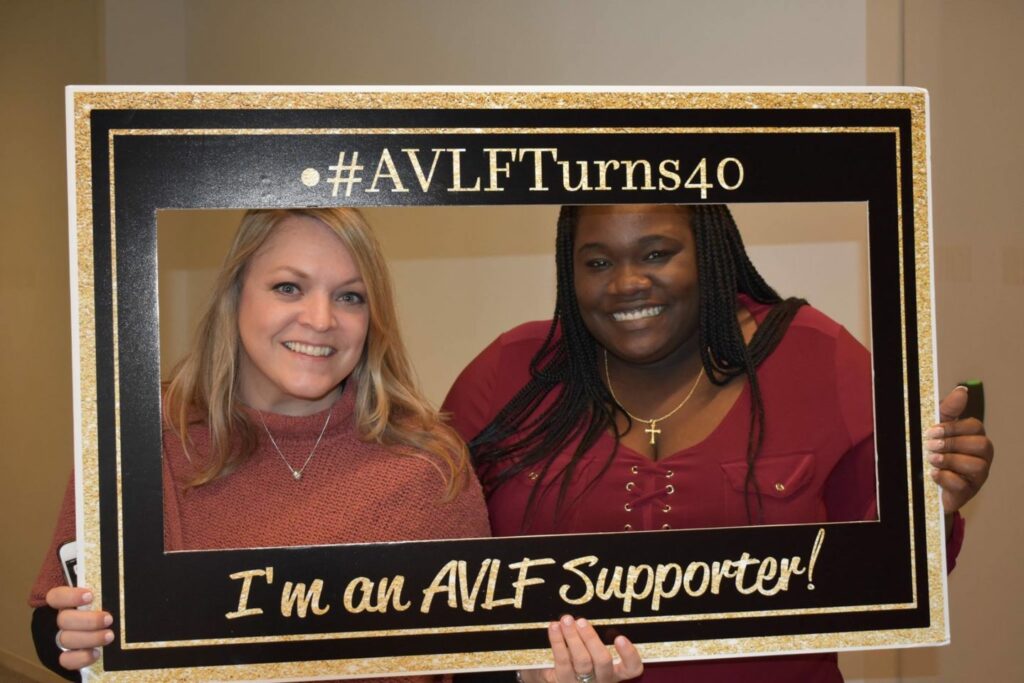 Two women smiling while holding a large black and gold frame with text: "#AVLFTurns40" and "I'm an AVLF Supporter!" The women appear to be at an indoor event celebrating AVLF's 40th anniversary.