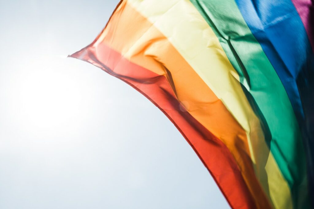 Close-up of a rainbow flag waving against a bright, blue sky. Sunlight shines through the fabric, highlighting the vibrant colors of red, orange, yellow, green, blue, and purple.