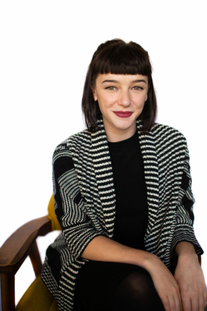 A person with short dark hair and a nose piercing sits on a chair. They are wearing a black top and a black-and-white striped cardigan. The background is plain white, and the person is smiling at the camera.