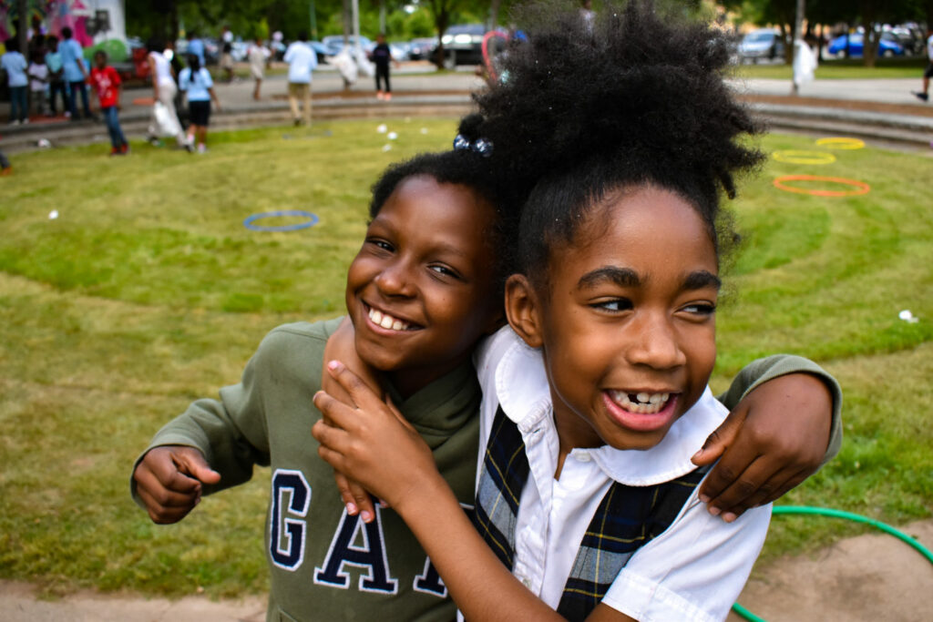 Two smiling children with arms around each other, standing on a grassy lawn. The background shows a few people, circular games, and a mural. The sky is clear, suggesting a sunny day.