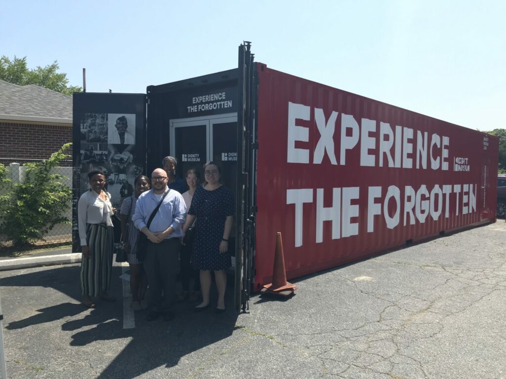 A group of five people stands smiling in front of a large red shipping container. The container has "EXPERIENCE THE FORGOTTEN" written in bold white letters. The scene is in an outdoor setting with a building and greenery in the background.