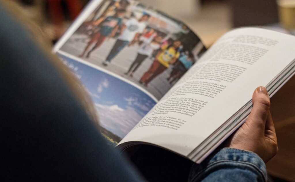 A person in a blue denim shirt is holding an open book. The left page features a photo of people walking, while the right page contains text. The background is blurred.