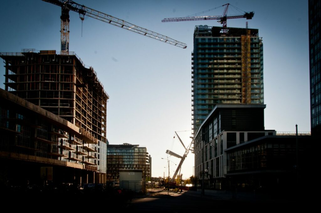 A city skyline at sunset with cranes towering over partially constructed high-rise buildings. Streets are deserted, and the sky has a warm, golden hue, highlighting the silhouettes of the cranes and buildings.