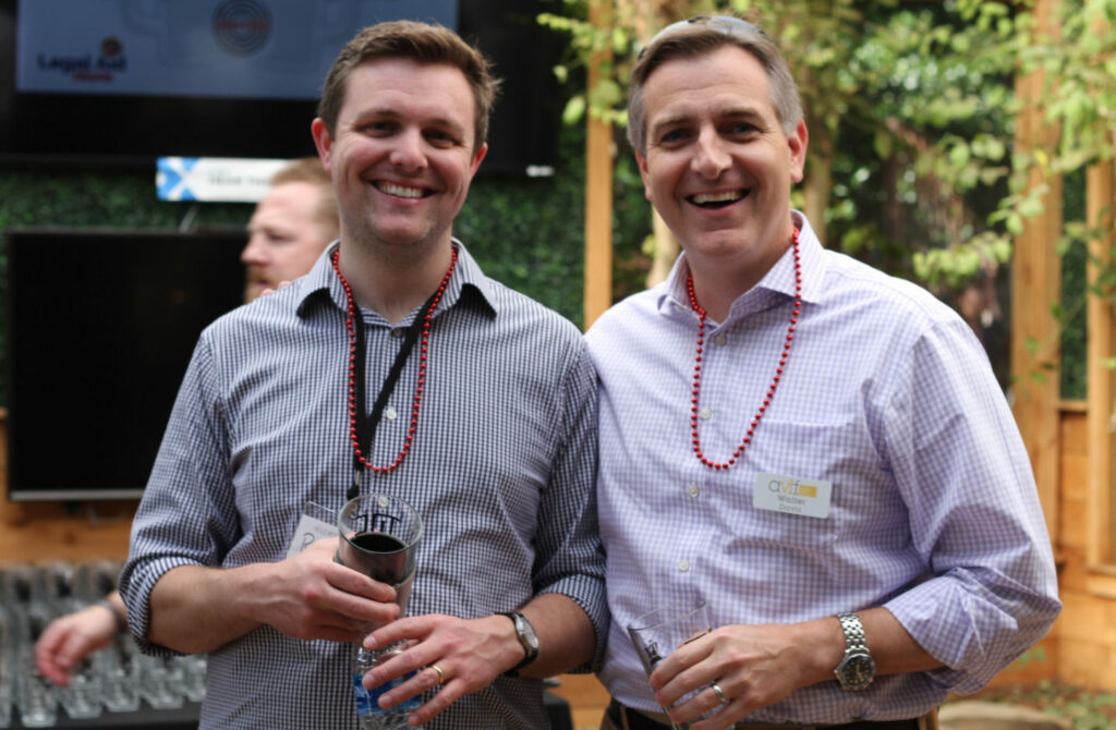 Two men smiling at the camera outdoors, wearing checkered shirts and red beaded necklaces. They are holding drinks, and there's greenery and a wooden structure in the background. Another person is slightly visible behind them.