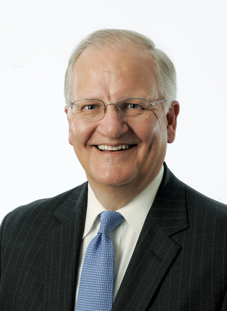 A middle-aged man smiling, wearing glasses, a dark pinstripe suit, white shirt, and a blue patterned tie, against a plain white background.