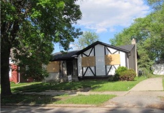 A small house with a Tudor-style facade has boarded-up windows. The surrounding area features a few trees, grass, and a concrete driveway under a clear blue sky.