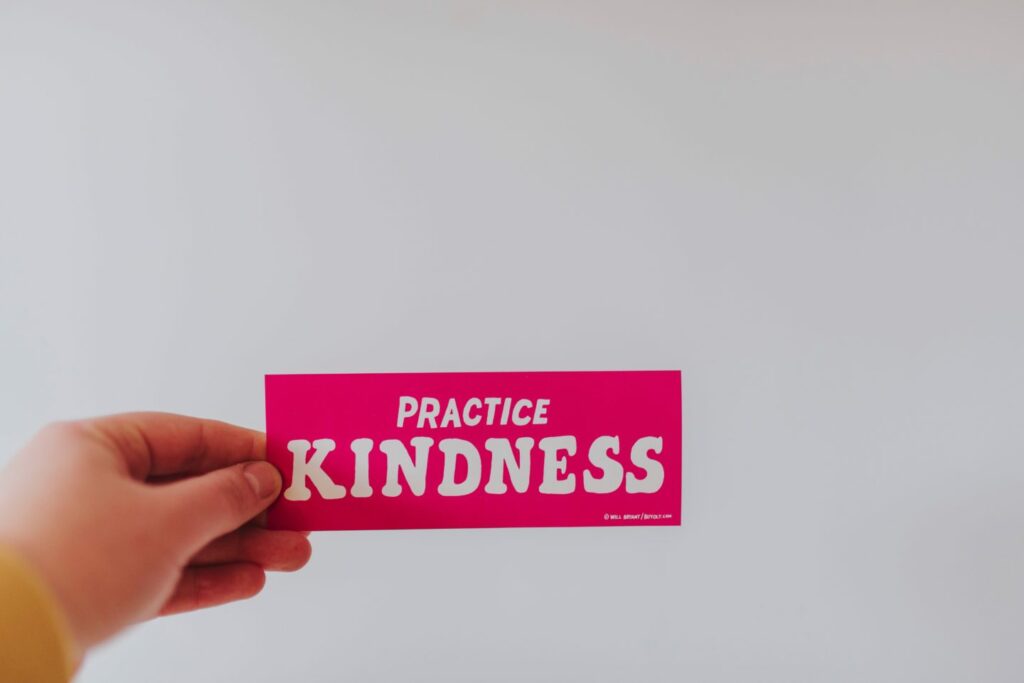 A hand holds a bright pink card with the message "Practice Kindness" in bold white letters against a plain light gray background.