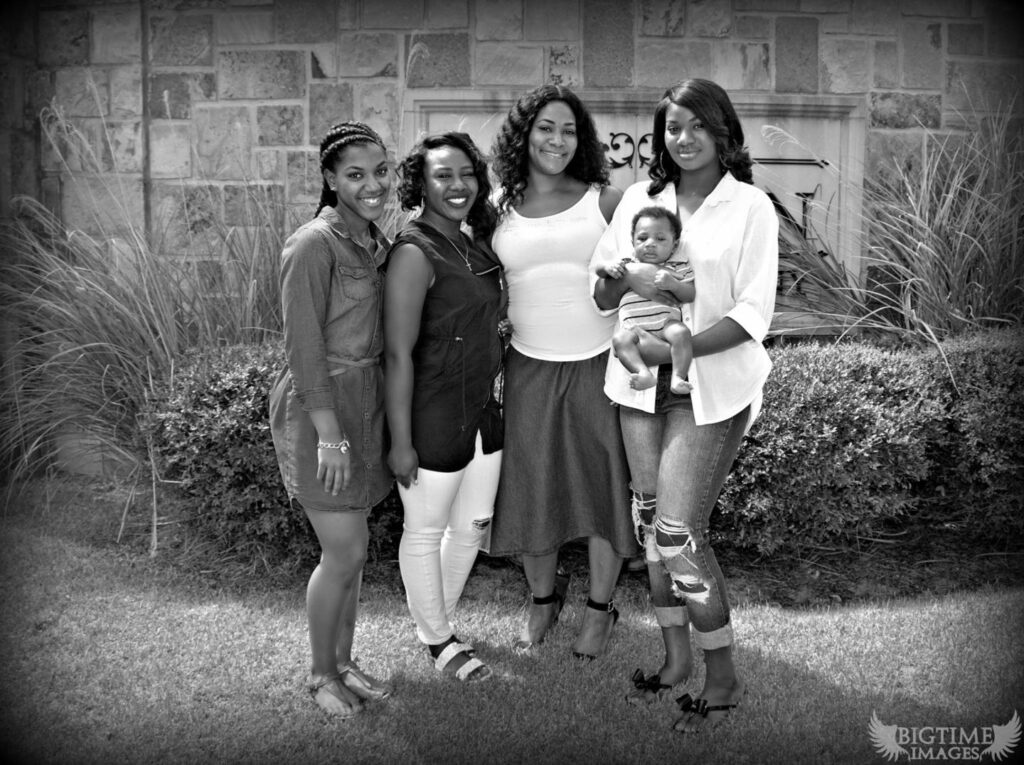A group of four women and a baby pose in front of a stone wall. Three women are standing, one woman in the center is barefoot, and another holds the baby. The setting is outdoors, with grass and plants surrounding them. The photo is in black and white.