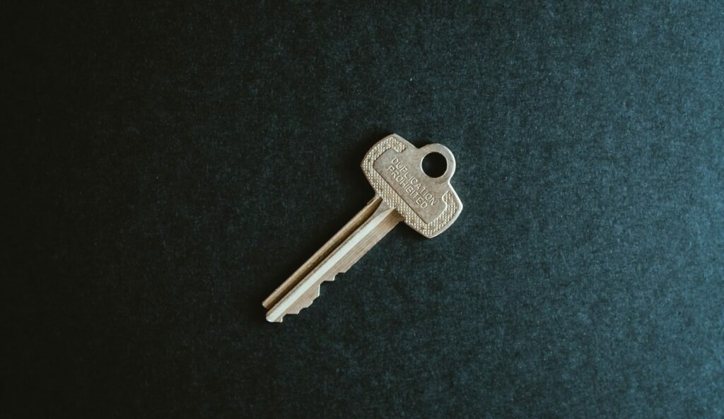 A single brass key with serrated edges lies flat on a textured dark surface. The key has a small round hole at the top for attachment and subtle engravings are visible. The overall setting is simple and minimalist.