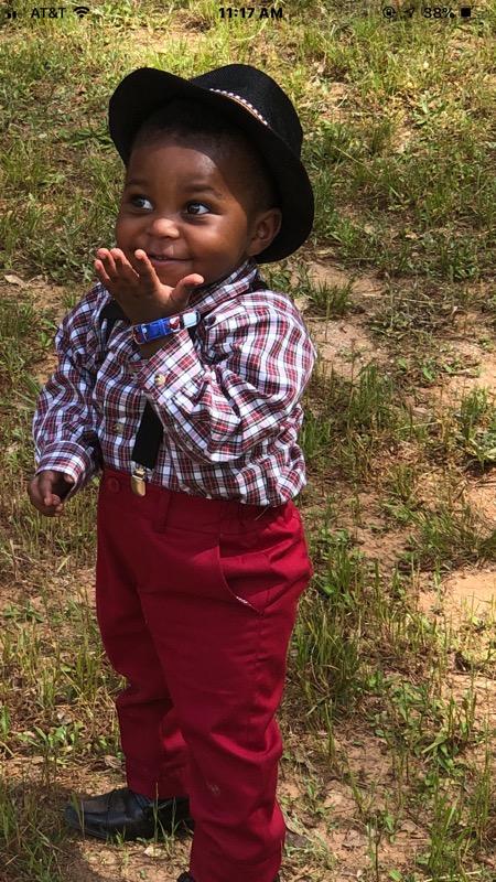 A young child stands outdoors on grass, wearing a black hat, a plaid shirt with suspenders, red pants, and black shoes. The child is smiling with one hand covering their mouth, looking to the side with an expression of delight.