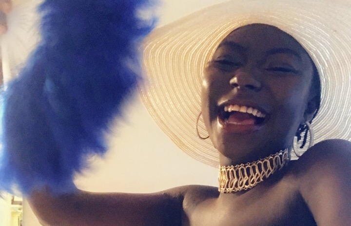 A person smiling joyfully while wearing a wide-brimmed hat, large hoop earrings, and a gold chain necklace. They are holding a blue feather accessory, adding a playful and vibrant element to the scene.