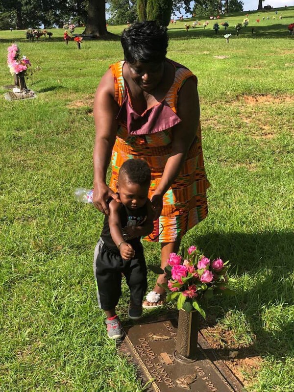 A woman and a young child stand together at a gravesite. The woman holds the child as they look at a headstone adorned with pink flowers. The setting is a grassy cemetery with other headstones and floral arrangements visible in the background.