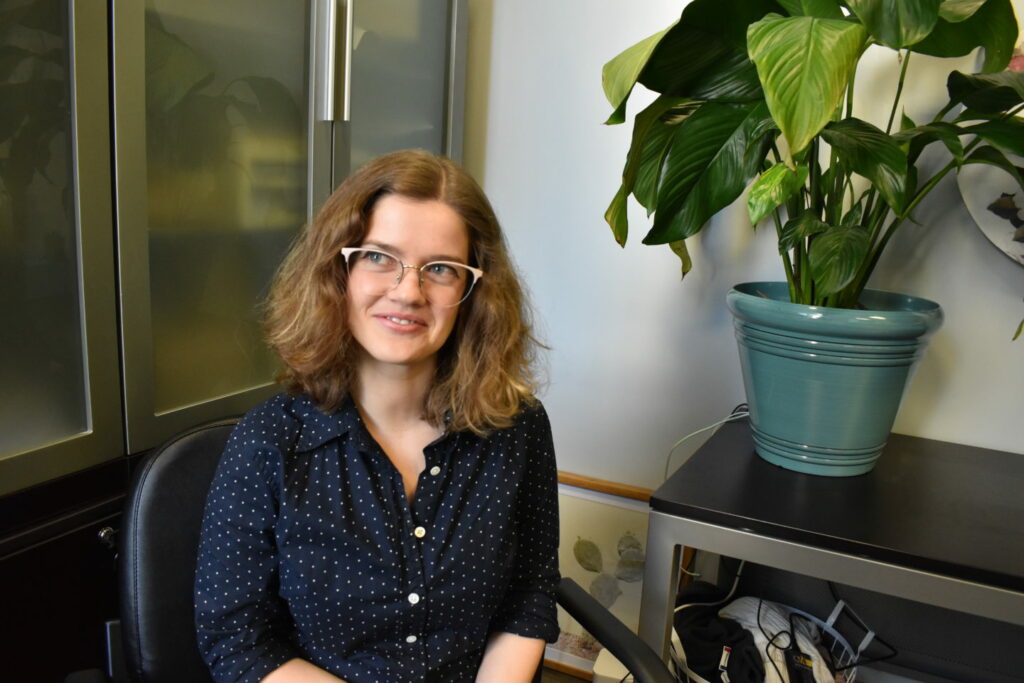 A person with wavy hair and glasses, wearing a navy blue polka dot shirt, smiles while sitting in an office chair. A large green plant in a blue pot is on a cabinet beside them.
