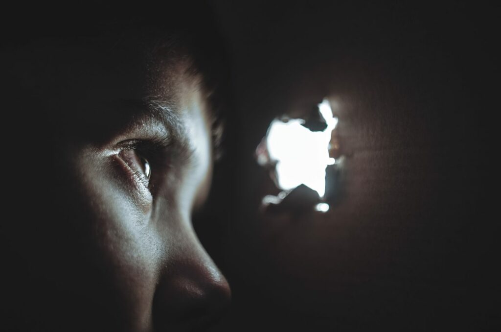 A person peers through a small hole in a dark surface, with light illuminating their eye and part of their face. The rest of the image is in deep shadow, creating a dramatic contrast between light and dark.