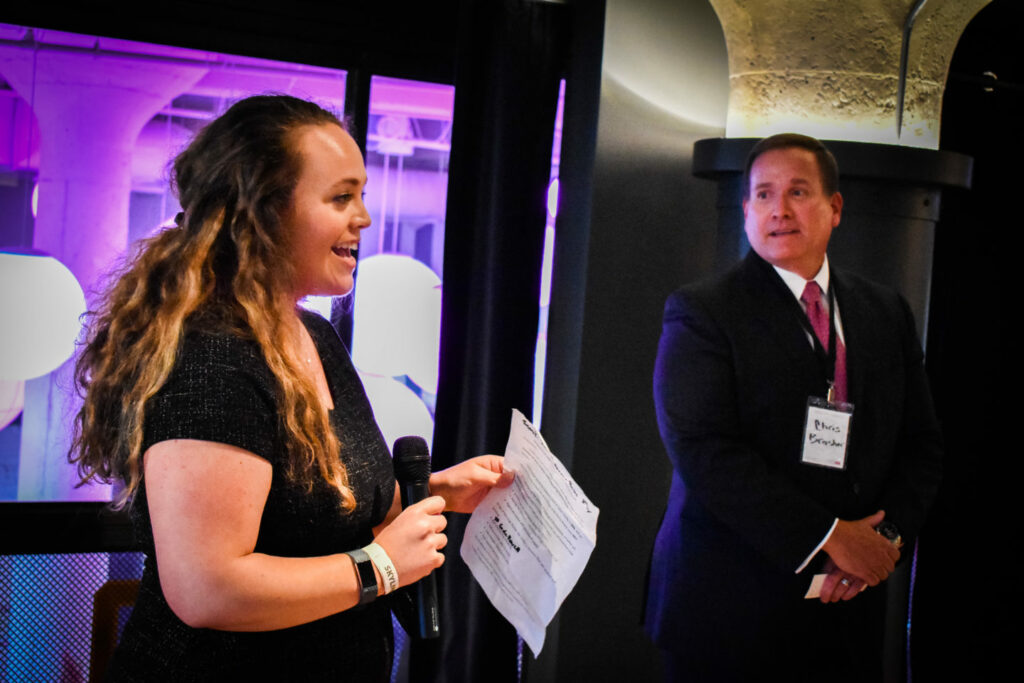 A woman speaks into a microphone while holding a paper, and a man in a suit with a name tag listens intently. The background is softly lit in purple hues.