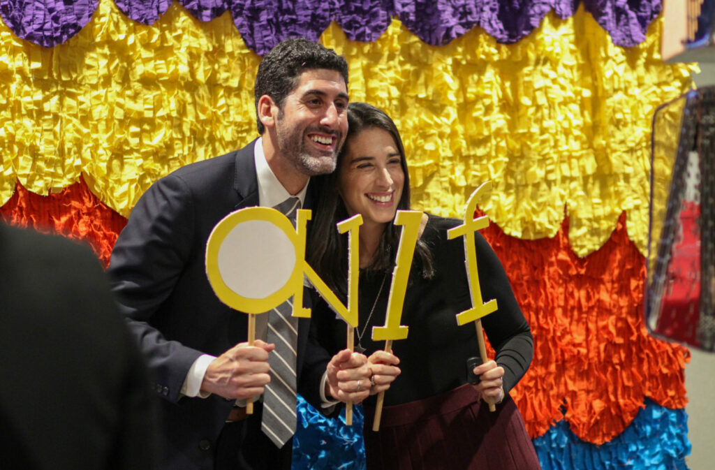 A man and woman smiling, holding yellow letter cutouts against a colorful backdrop with yellow, purple, and orange fringes. The letters spell "CNMF." They appear to be at a festive event.