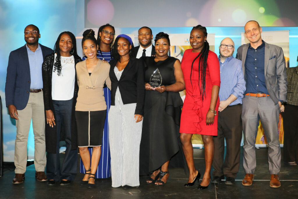 A diverse group of eleven people stands together on a stage. One person holds an award. They are all smiling and dressed in semi-formal attire, with a colorful backdrop behind them.