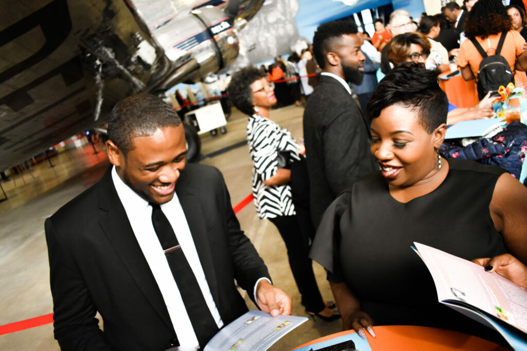 A man and woman smiling while reading a booklet at an indoor event. They appear to be under a large aircraft, with other people socializing in the background. The setting is lively with a mix of formal and festive attire.