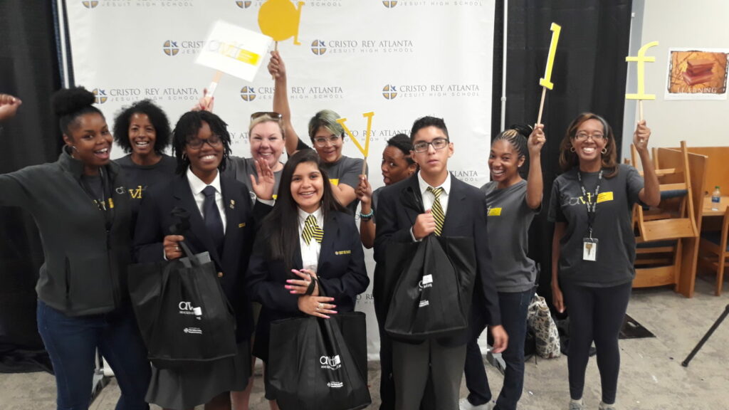 A group of smiling students and staff pose together at Cristo Rey Atlanta Jesuit High School. Some hold signs with messages and logos, while others carry gift bags, standing in front of a branded backdrop.