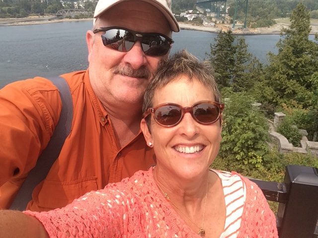 A man and a woman smiling and taking a selfie on a sunny day. The man is wearing a cap and orange shirt, and the woman is wearing sunglasses and a peach-colored top. They are standing near a body of water with trees and a bridge in the background.