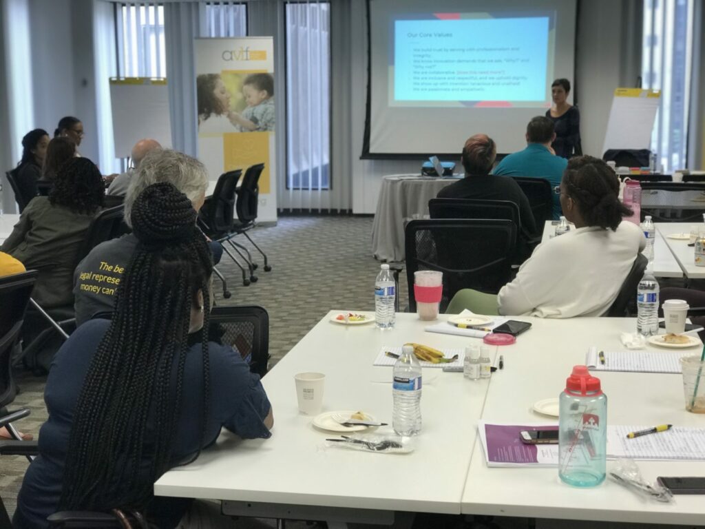A group of people seated at tables in a conference room, listening to a presentation. A woman stands near a screen displaying a slide titled "Our Core Values." Notebooks, cups, and snacks are on the tables.