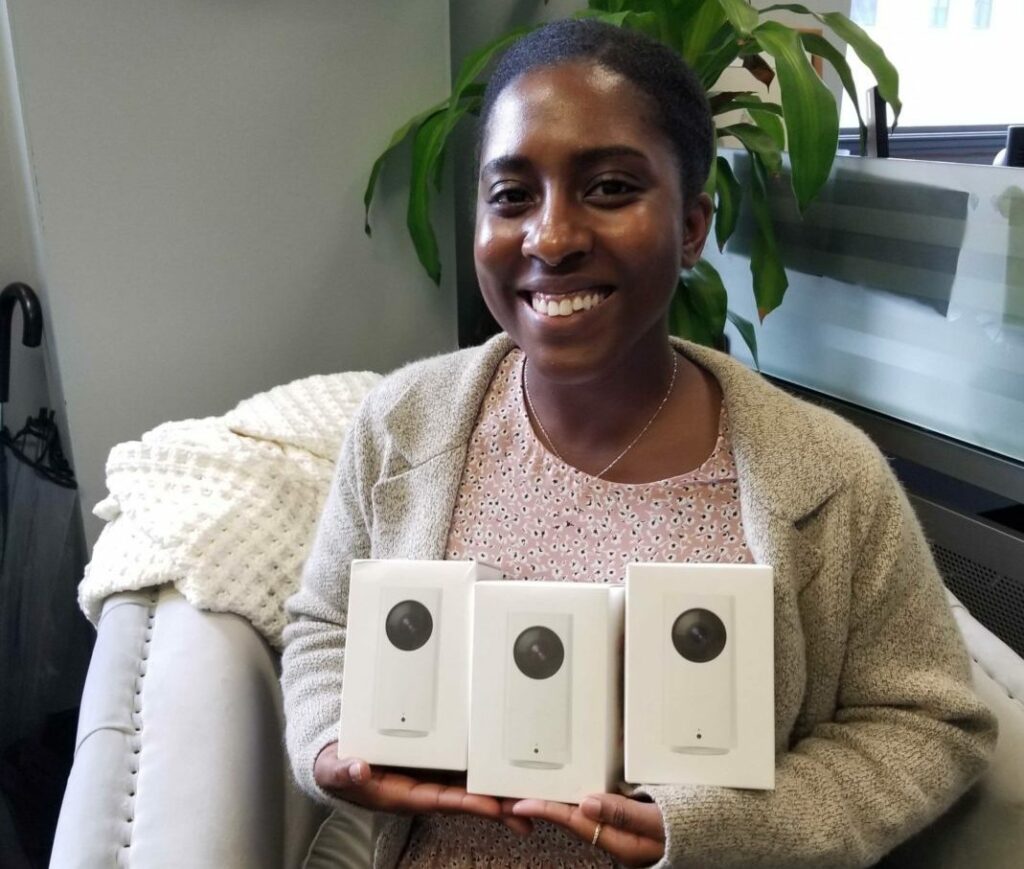A person smiling while holding three white boxes with electronic devices inside, sitting on a sofa with a blanket nearby. There's a plant in the background.