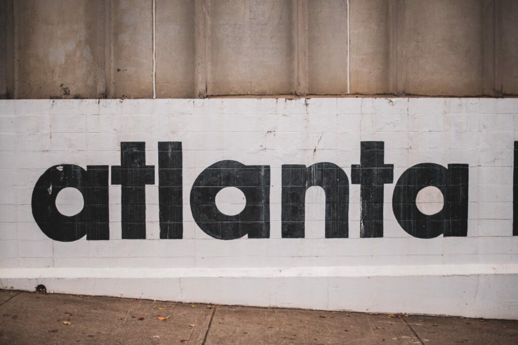 The image shows a concrete wall with the word "atlanta" painted in large black letters. The wall and sidewalk in front are slightly worn, with a few scattered leaves on the ground.