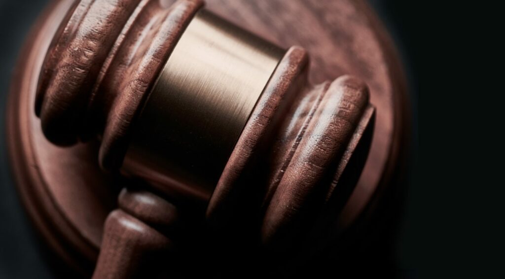Close-up of a wooden gavel resting on a sound block against a dark background, evoking the question, "Where's home?" The gavel shows rich texture and detail, emphasizing its polished wood finish and metal band around the head.