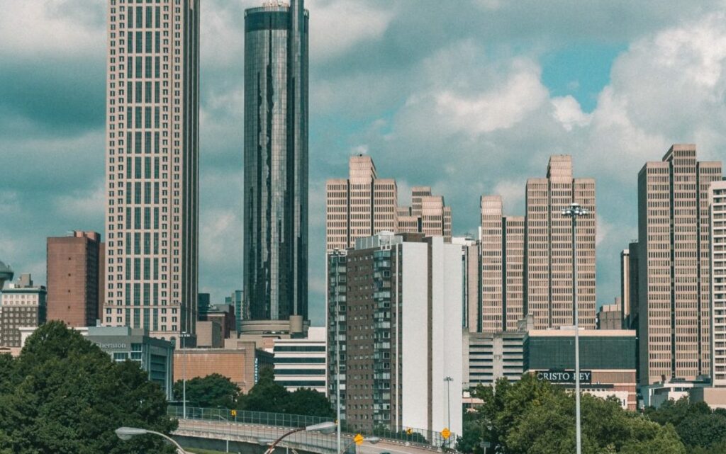 A cityscape featuring tall skyscrapers and modern high-rise buildings under a partly cloudy sky. Trees and urban infrastructure, including roads and signs, are visible in the foreground.