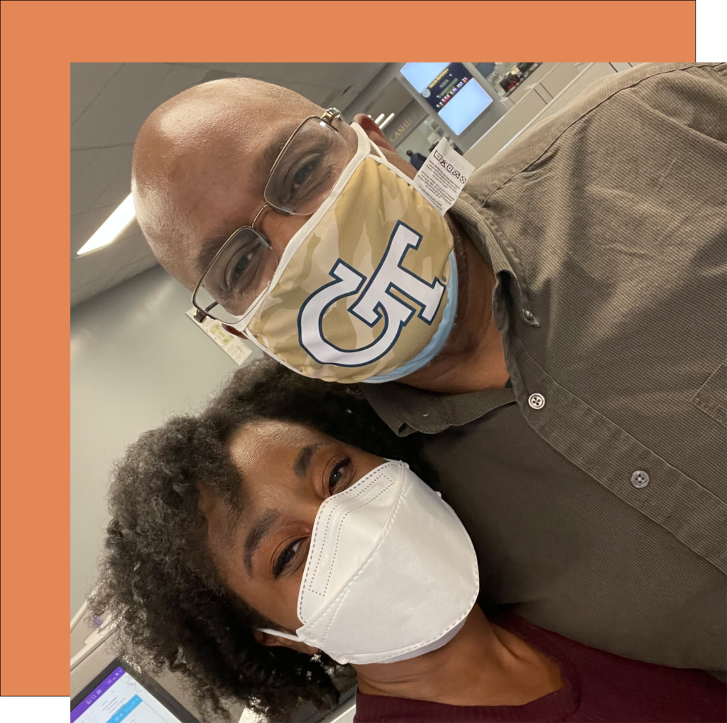 A man and a woman wearing face masks stand close together and smile for a selfie. The man wears glasses and a mask with a logo, while the woman wears a plain white mask. They are indoors, with a computer workstation visible in the background.