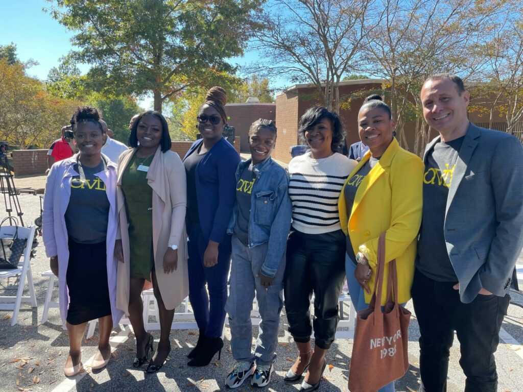 A group of seven smiling people stand outdoors in front of trees and buildings, some wearing "CWDI" shirts. They are casually dressed, with jackets and coats, and appear to be enjoying a sunny day.