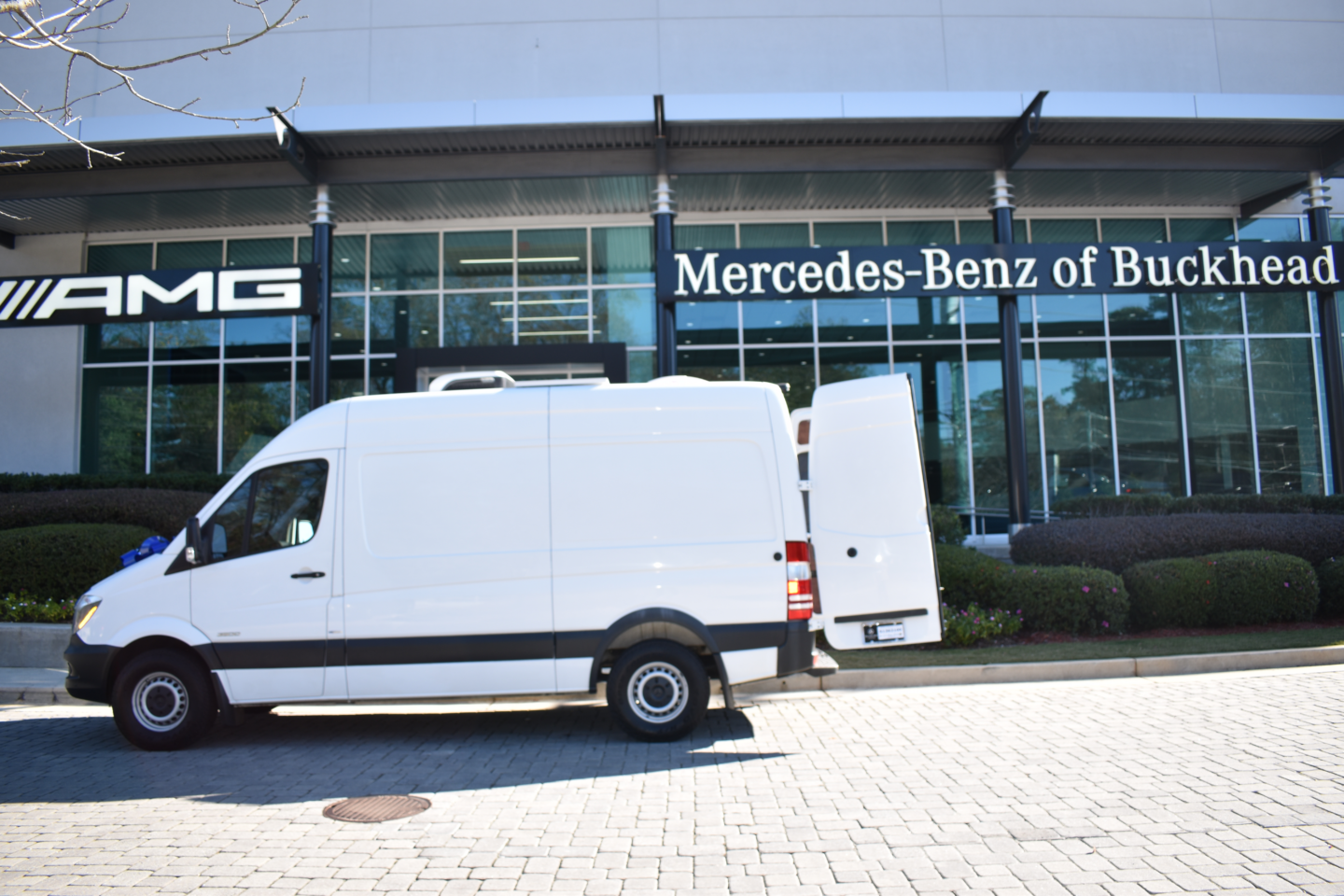 A white delivery van is parked outside the Mercedes-Benz of Buckhead dealership. The rear door of the van is open, and the dealership's building features large glass windows and a sign with the Mercedes-Benz logo.
