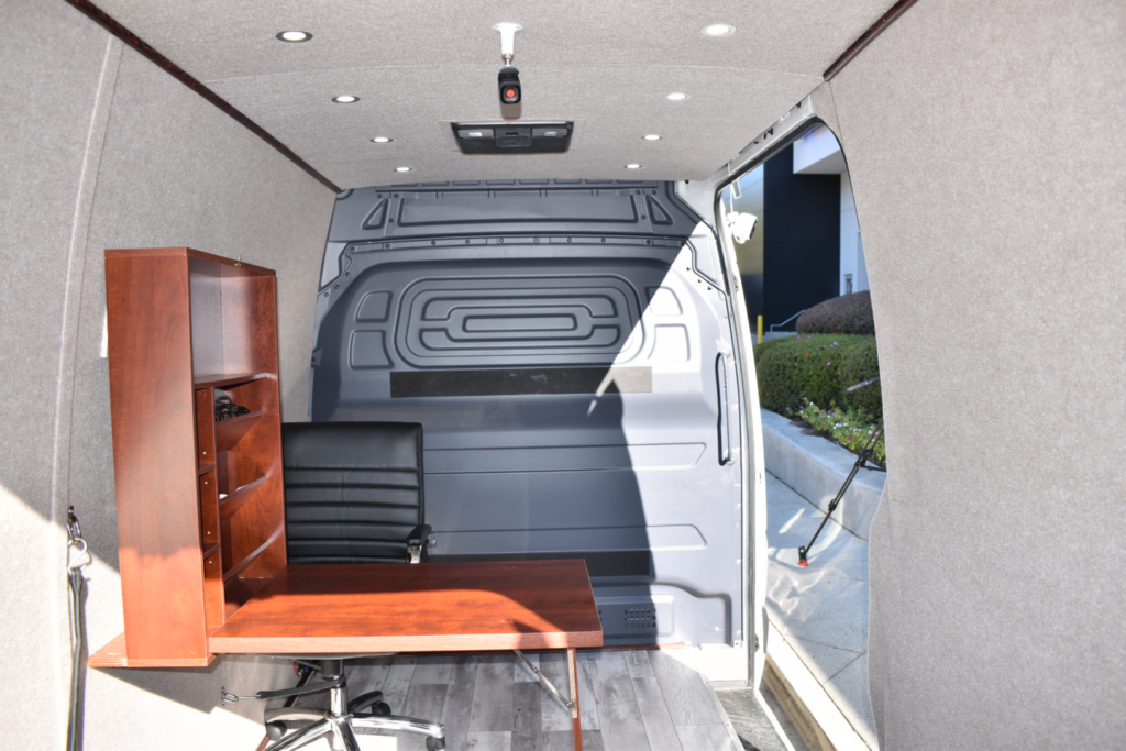 Interior of a van converted into an office space, featuring a wooden desk with an office chair and a shelf unit. The van has gray walls and a wood-like floor with sunlight coming through the open side door.