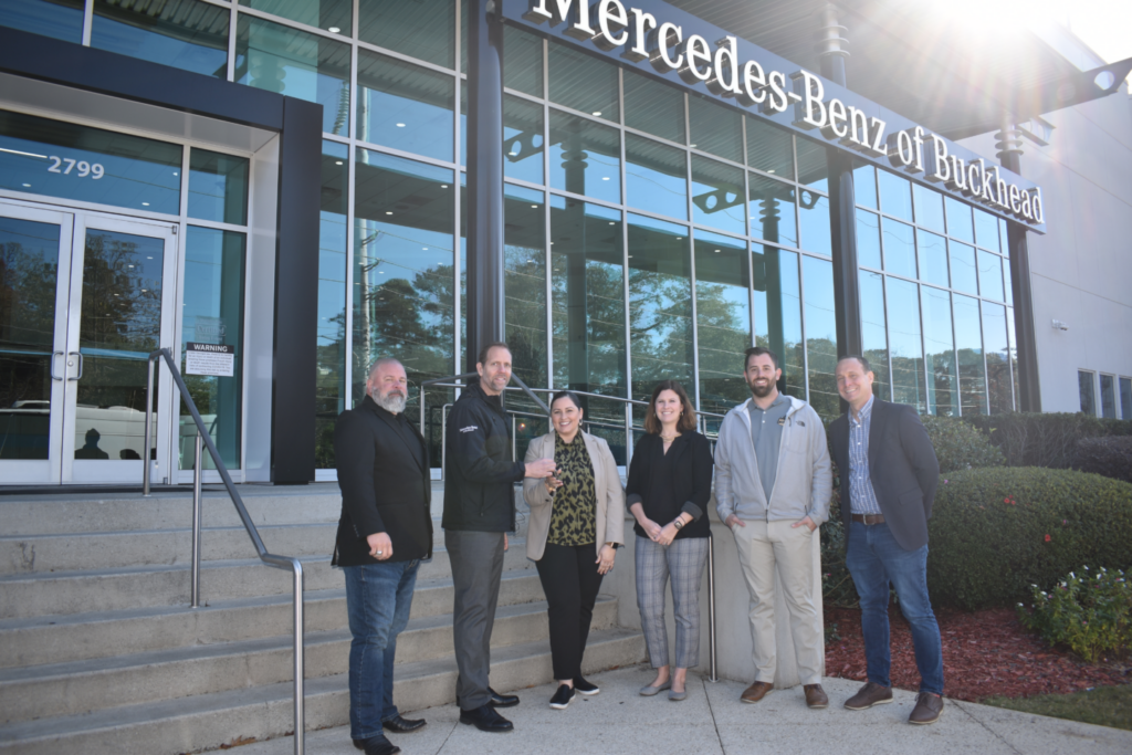 A group of six people stand in front of the entrance to Mercedes-Benz of Buckhead. The dealership building features large glass windows and the company’s logo prominently displayed above.