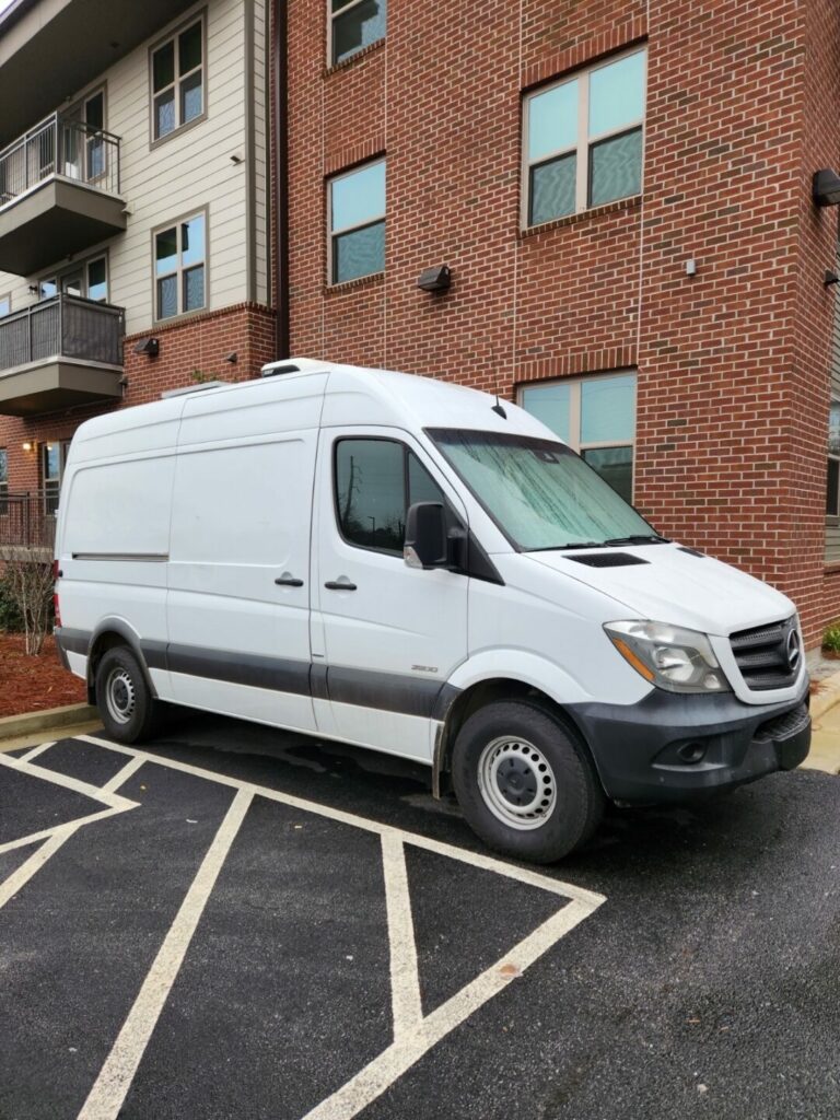 A white cargo van is parked in a designated parking space next to a brick building with multiple windows. The building has balconies, and the surrounding area includes a sidewalk and some landscaping.