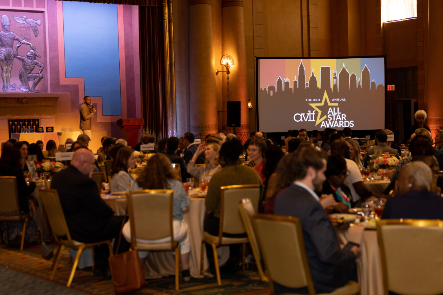 A large event with people seated at round tables, attentively listening to a speaker on stage. A screen displays "3rd Annual All Star Awards" with a city skyline graphic. The venue has warm lighting and elegant decor.