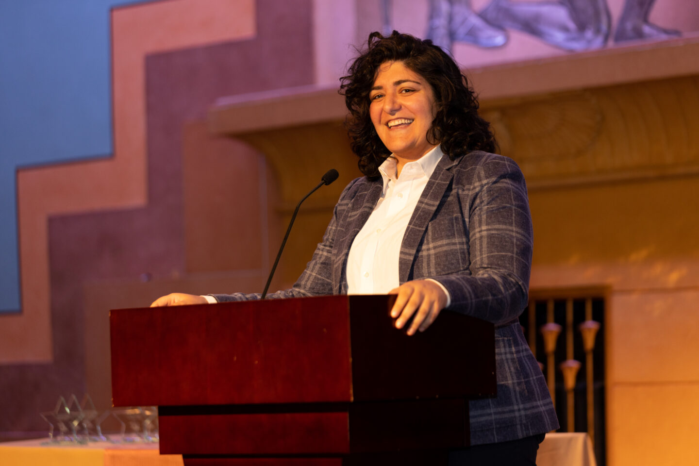 A person with curly hair stands at a podium, smiling and speaking into a microphone. They are wearing a plaid blazer and white shirt. The background features a warm-toned wall with artistic designs.