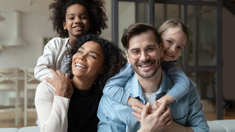 A joyful family scene with two smiling parents seated closely, each carrying a laughing child on their back. They are indoors, surrounded by a warm, cozy atmosphere. The family radiates happiness and togetherness.